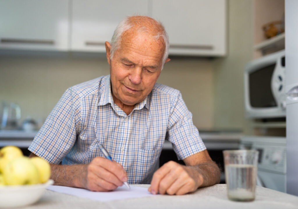 Senior man writing letter on paper while sitting in kitchen at home